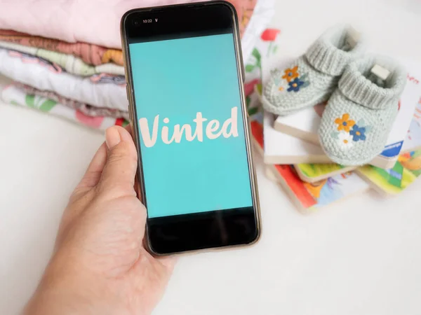 Braunschweig, Almanya - Juli 03, 2023: Elinde mobil uygulaması Vinted ve çocuk kıyafetleri olan bir akıllı telefon, beyaz arkaplan üzerine kitaplar. İnternet üzerinden çocukların ikinci el giysilerinin satışı
