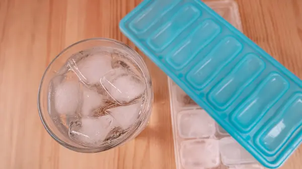 stock image Sparkling water with bubbles in a big transparent glass filled with ice cubes and a soft plastic blue form to make ice cubes in the freezer. Solution to overcome extreme high temperature in summer