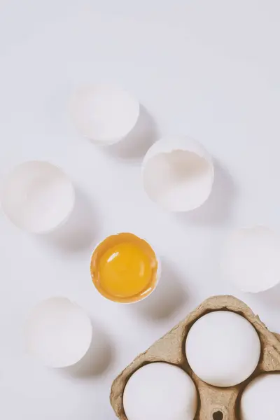 stock image Cracked egg half with a yolk inside near the egg tray with eggs on bright background. Selective focus. Concept scene. Top view