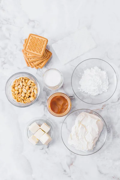 stock image Ingredients for cheesecake. Cookies, caramel, peanuts, butter, cream cheese, powdered sugar, cream in glassware and gelatin sheets on table. Top view.