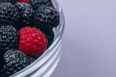 Gri arka planda kırmızı ve siyah ahududu olan cam kaseye yakın plan. Makro çekim.