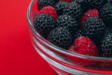 Close-up of glass bowl with red and black raspberries on red background. Macro shot.