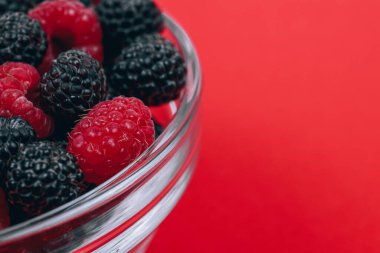 Close-up of glass bowl with red and black raspberries on red background. Macro shot.