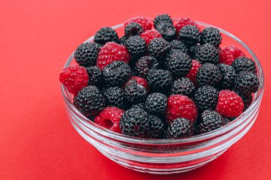 Close-up of glass bowl with red and black raspberries on red background. Macro shot.