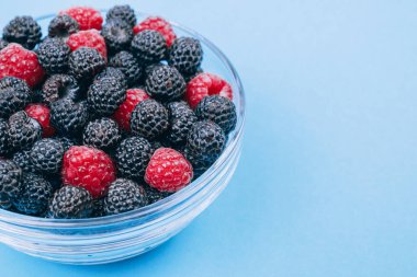 Mavi arka planda kırmızı ve siyah ahududu olan cam kaseye yakın plan. Makro çekim.