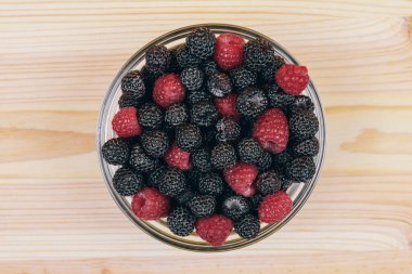 Ahşap masada kırmızı ve siyah ahududu olan cam kasenin en üst görüntüsü. Makro çekim.