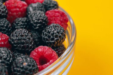 Sarı arka planda kırmızı ve siyah ahududu olan cam kaseye yakın plan. Makro çekim.