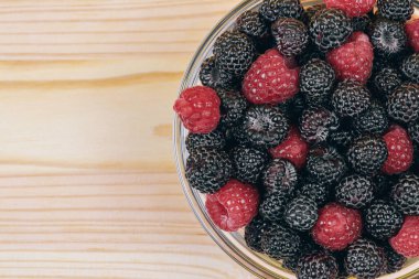 Ahşap masada kırmızı ve siyah ahududu olan cam kasenin en üst görüntüsü. Makro çekim.