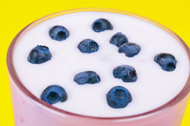 Glass of cold milkshake with fresh blueberries on top. Drink in a glass with a straw on a yellow background.