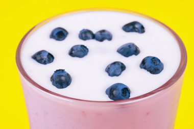 Glass of cold milkshake with fresh blueberries on top. Drink in a glass with a straw on a yellow background.