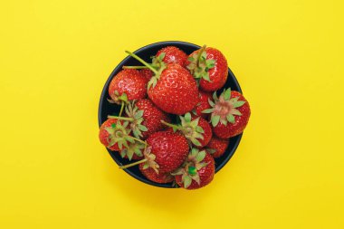 Olgun çilek. Sarı arka planda çilekli siyah kase. Üst görünüm.