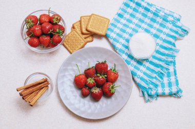Hafif bir masada toplanmış taze çilek yığını. Seramik tabakta çilek ve bir kase şeker, tarçın ve kurabiyeyle servis edilen cam kase. Üst görünüm.