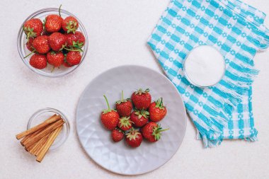 Hafif bir masada toplanmış taze çilek yığını. Seramik tabakta çilek ve bir kase şeker ve tarçınlı cam kase. Üst görünüm.