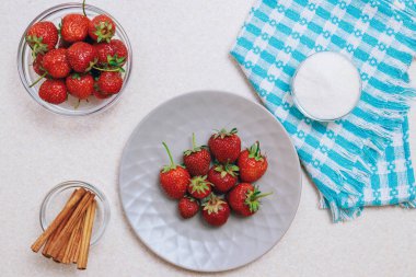 Hafif bir masada toplanmış taze çilek yığını. Seramik tabakta çilek ve bir kase şeker ve tarçınlı cam kase. Üst görünüm.