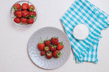 Hafif bir masada toplanmış taze çilek yığını. Seramik tabakta çilek ve bir kase şekerle birlikte cam kase. Üst görünüm.