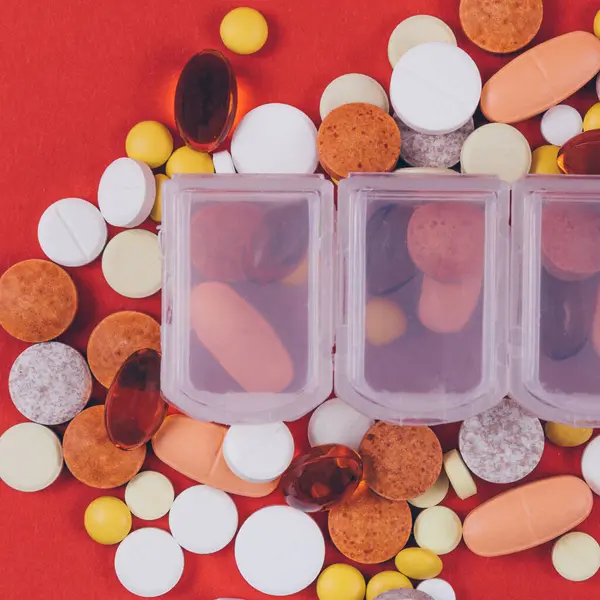 Multicolor pills and tablets in and out of pill organizer on red background. Various treatments mixed. Variety of pills and medication. Close up. Macro. Medical concept. Top view.