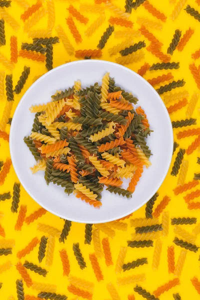 Three-colored uncooked pasta in the white bowl on yellow background with scattered pasta on it. Top view.