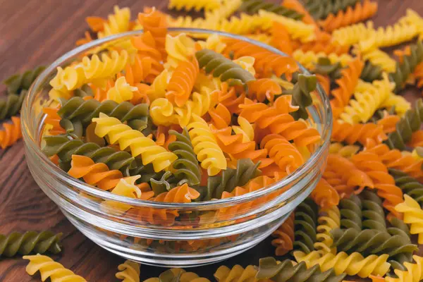 Three-colored uncooked pasta in the glass bowl on wooden background with scattered pasta on it.
