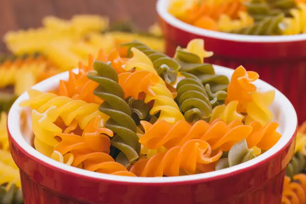 Three-colored uncooked pasta in the ceramic bowls on the wooden background with scattered pasta on it.