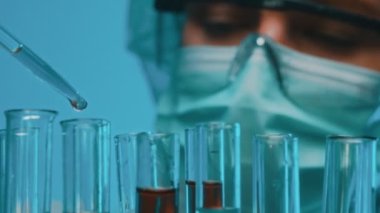 Gözlüklü ve maskeli bir kadın laboratuarda test tüpleriyle çalışıyor..