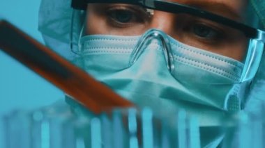 Gözlüklü ve maskeli bir kadın laboratuarda test tüpleriyle çalışıyor..