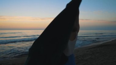 Woman throws a blanket over herself at sunset by the sea.