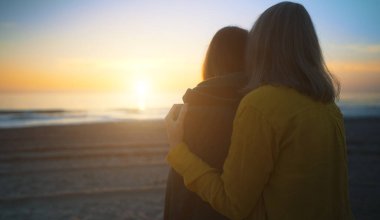 Mother cover his daughter with blanket and hugging on the beach at sunset.