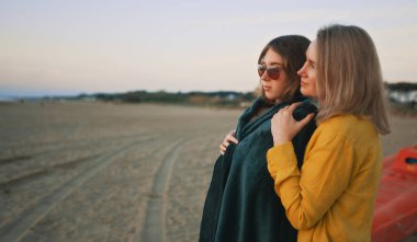 Mother cover his daughter with blanket and hugging on the beach at sunset.