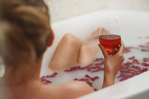 Woman enjoys wine in a bath with milk and rose petals.