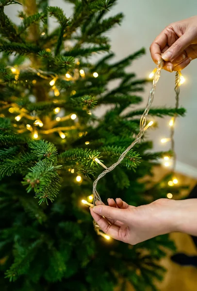 Woman hangs garland with light bulbs on a New Year tree.