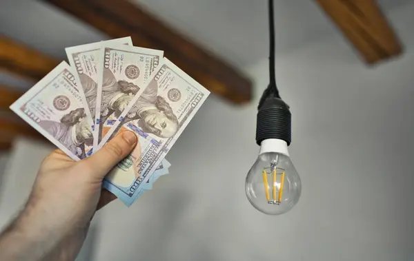 Man holds money next to a light bulb. Expensive electricity concept.