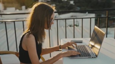 Genç kız gün batımında balkonda dizüstü bilgisayarda çalışıyor..