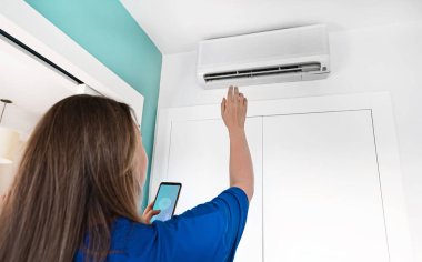 Telefonundaki uzaktan kumandayla akıllı bir klimayı kontrol eden bir kadın..