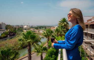 Kırmızı şaraplı mavi elbiseli kadın balkonda tatilin tadını çıkarıyor..