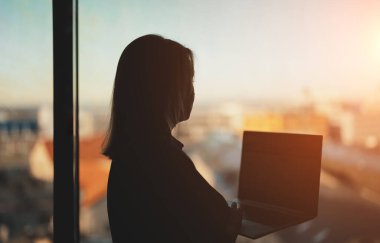 Ofis penceresinin yanında dizüstü bilgisayarı olan bir kadının silueti..