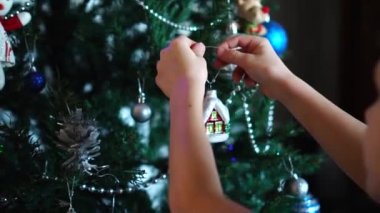 Yapay Noel ağacını ev şeklinde bir Noel oyuncağıyla süsleyen bir çocuğun elleri. Tatil için hazırlık atmosferi.