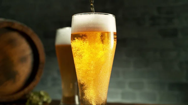 Glass of light beer pouring on wooden table. Still life shot with wooden keg on background.