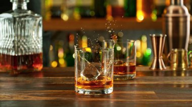 Ice cube fall into a glass with a golden alcoholic drink. Bar on background.
