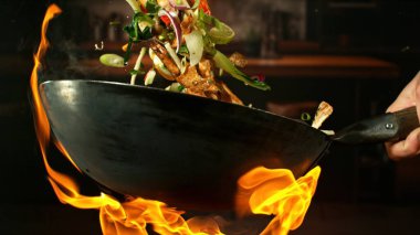 Wok Pan 'ın Dondurucu Hareketi ve Havadaki Uçan Malzemeler ve Ateş Alevleri. Arka planda Mutfak İçi.