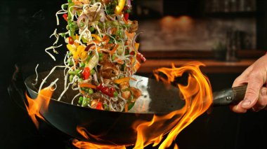 Wok Pan 'ın Dondurucu Hareketi ve Havadaki Uçan Malzemeler ve Ateş Alevleri. Arka planda Mutfak İçi.