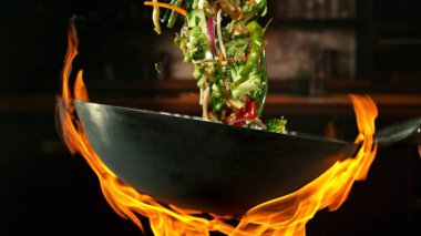 Wok Pan 'ın Donmuş Hareketi ve Havada Uçan Sebze İçindekiler ve Ateş Alevleri. Arka planda Mutfak İçi.