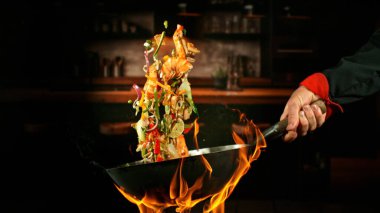 Wok Pan 'ın Dondurucu Hareketi ve Havadaki Uçan Malzemeler ve Ateş Alevleri. Arka planda Mutfak İçi.