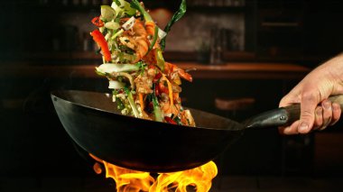 Wok Pan 'ın Dondurucu Hareketi ve Havadaki Uçan Malzemeler ve Ateş Alevleri. Arka planda Mutfak İçi.