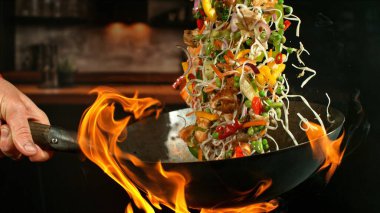 Wok Pan 'ın Dondurucu Hareketi ve Havadaki Uçan Malzemeler ve Ateş Alevleri. Arka planda Mutfak İçi.