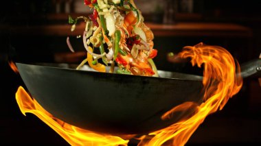 Wok Pan 'ın Dondurucu Hareketi ve Havadaki Uçan Malzemeler ve Ateş Alevleri. Arka planda Mutfak İçi.