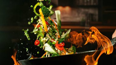 Wok Pan 'ın Donmuş Hareketi ve Havada Uçan Sebze İçindekiler ve Ateş Alevleri. Arka planda Mutfak İçi.