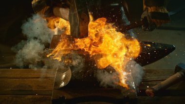 Demirci, kıvılcım saçan havai fişeklerle örsün üzerindeki erimiş metali elle dövüyor. Aksiyon çekimi, sinematik ve dramatik ışıklar. Hareketi dondur.