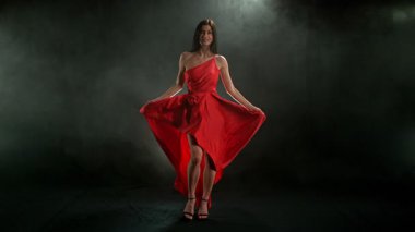 Yakut kırmızısı elbiseli, çarpıcı siyah saçlı kadın ve yüksek topuklu ayakkabı giyen zarif siyah arka plan pozu, dramatik ve zarif bir moda efekti için süper yavaş çekimde yakalandı..