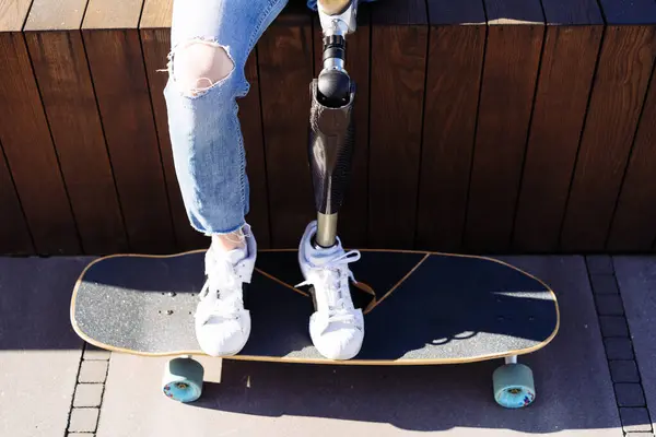 Close up of women prosthetic leg on skateboard in city. Disabled woman with prosthetic leg sitting on wood bench. Woman with leg prosthesis equipment.