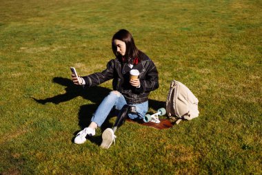 Protez bacaklı genç bir kız öğrenci üniversite kampüsünde yeşil çimlerde oturuyor ve akıllı telefonuyla selfie çekiyor. Biyonik bacağı olan engelli bir kadın. Telefon kullanan bacak protezi olan bir kadın..